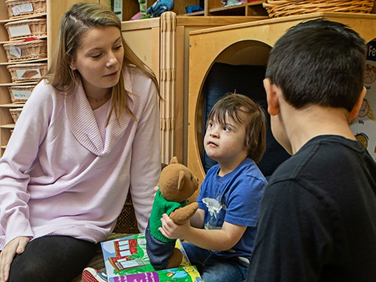An adult and two children sitting on the floor in a classroom area, with one child holding a stuffed bear and books spread out nearby.