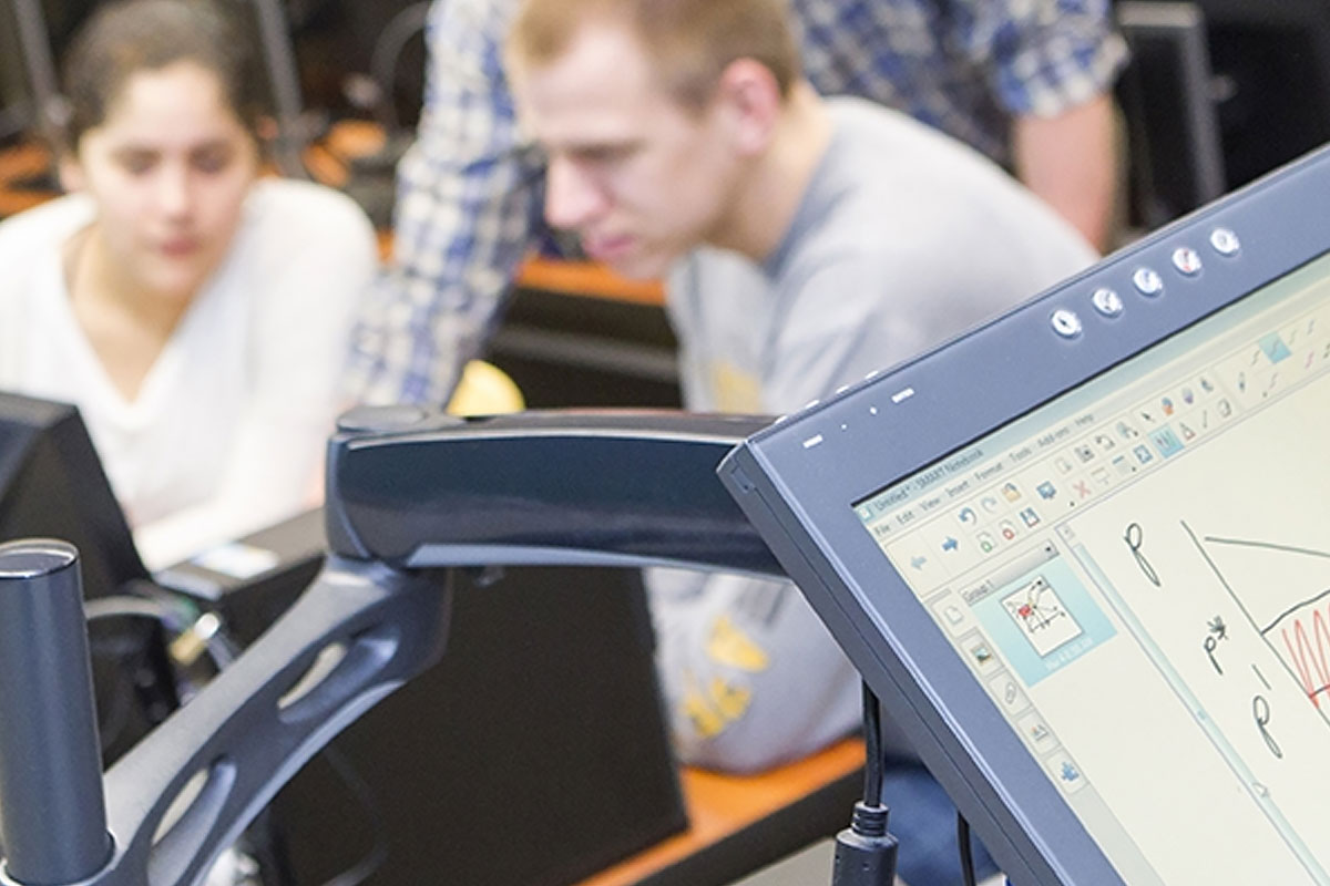 The primary focus is a monitor displaying a supply and demand graph with a student and an instructor reviewing material on a monitor..