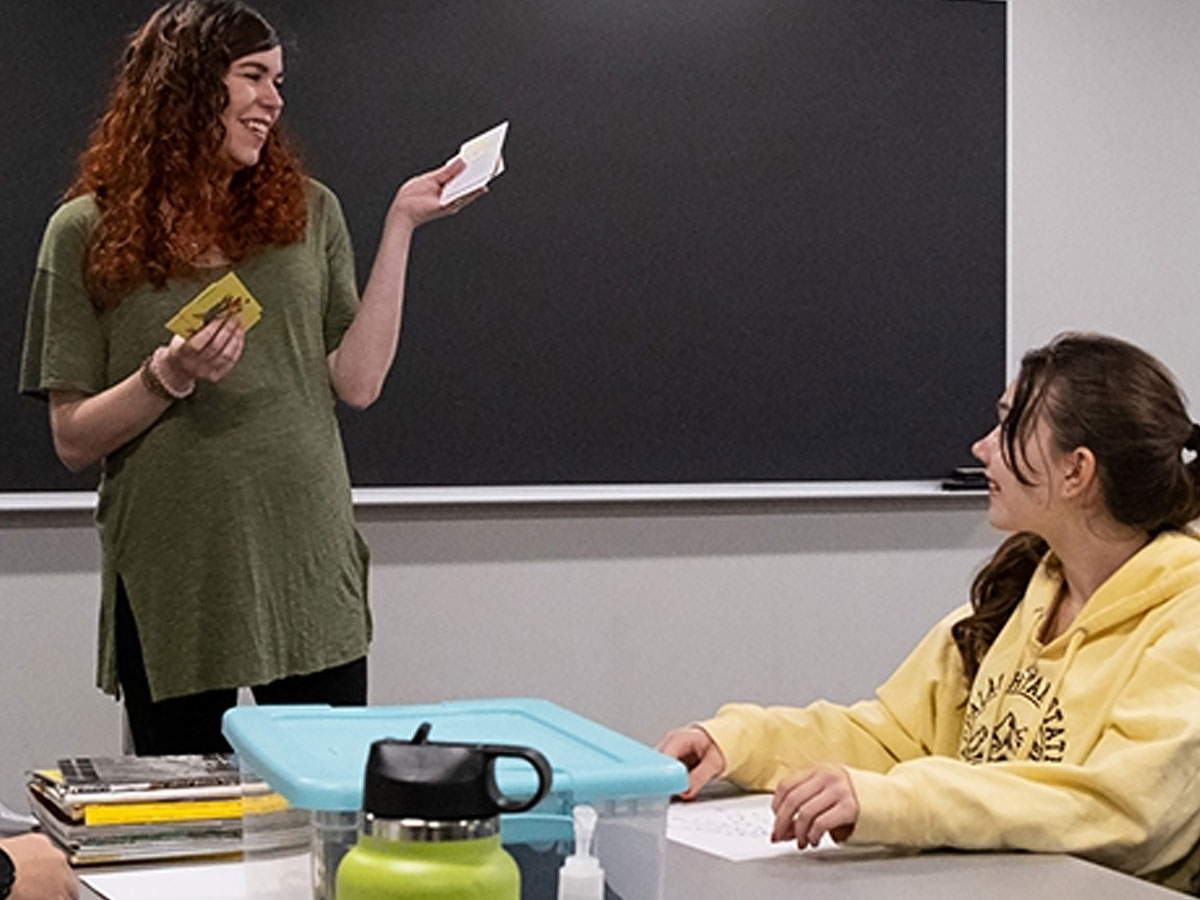 A person standing at the front of a classroom holding small cards, while another person sits at a table listening, with books and a storage bin on the desk.