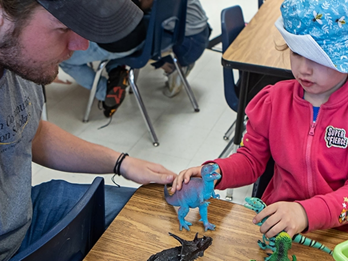 An adult and a child seated at a classroom table playing with plastic dinosaur toys, with others in background.