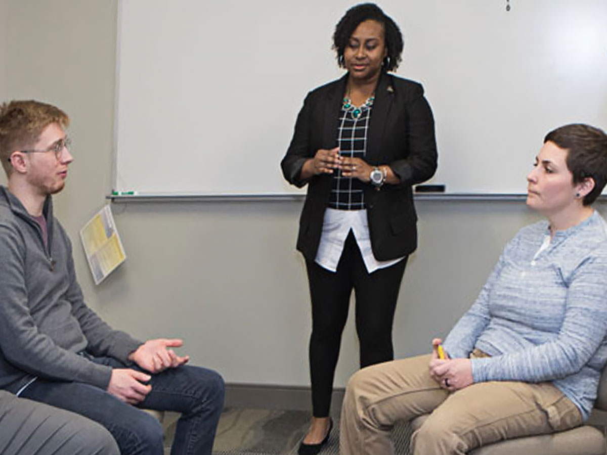 Three people in a classroom setting, with one standing near a whiteboard and two seated in chairs facing each other.