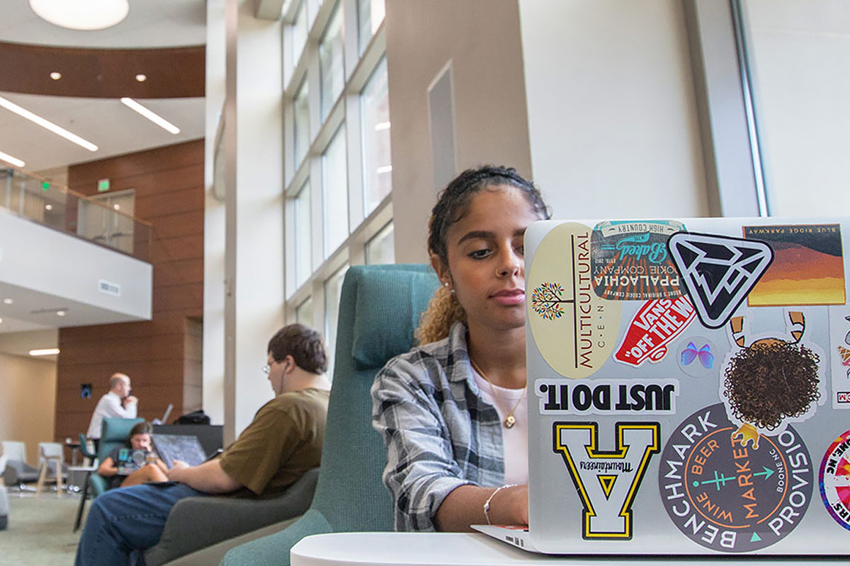 A student working on a laptop covered in stickers, including local Boone, NC references.