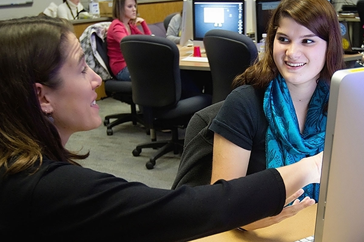 Two individuals collaborate at computer workstations in a classroom, with one person pointing at a screen.