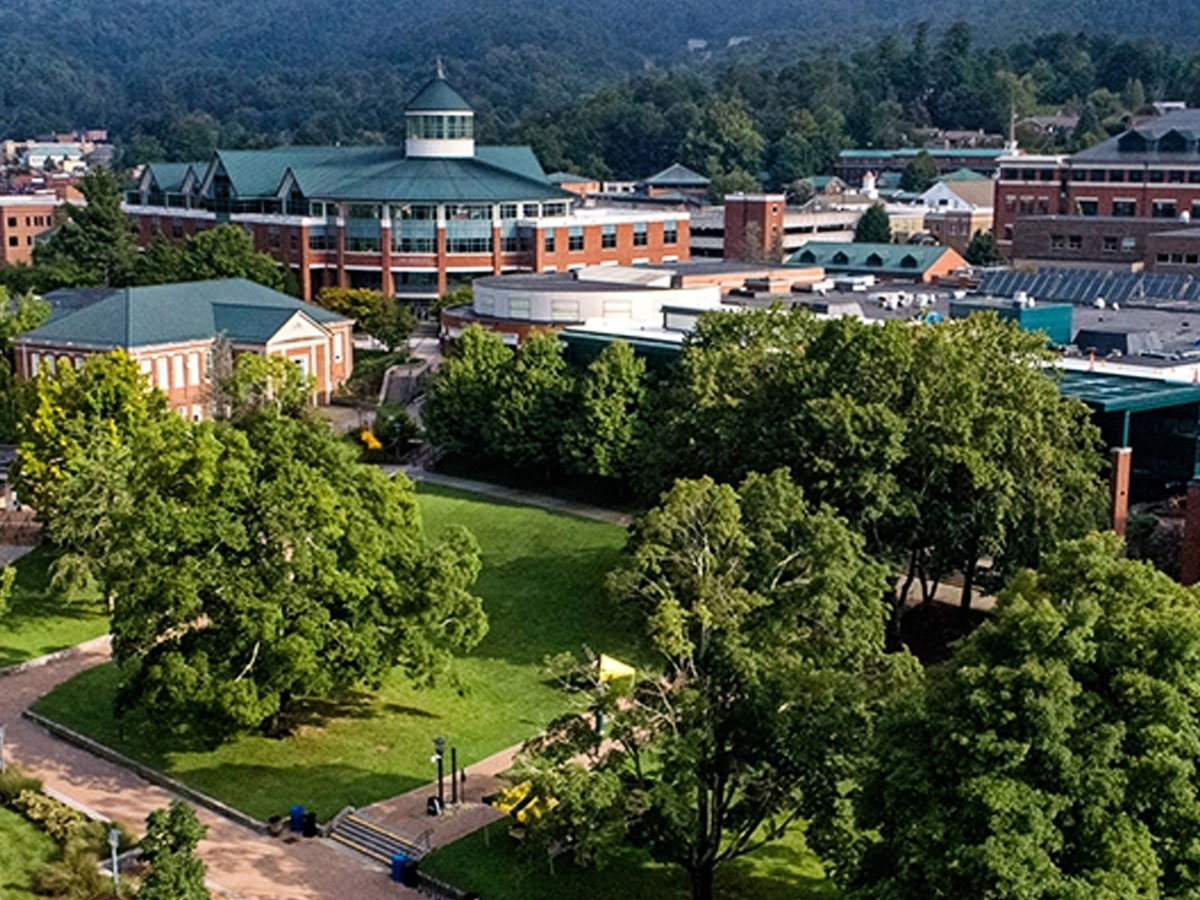 Aerial view of a App State campus with multiple brick buildings, large green spaces, and surrounding mountains.