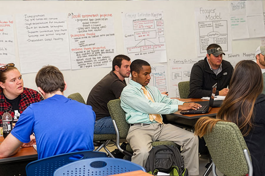 Adults seated at tables in a learning space, working on laptops and discussion activities.