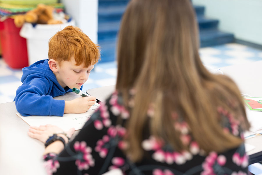 Child writing or drawing at a classroom table while an adult observes and supports learning activity.