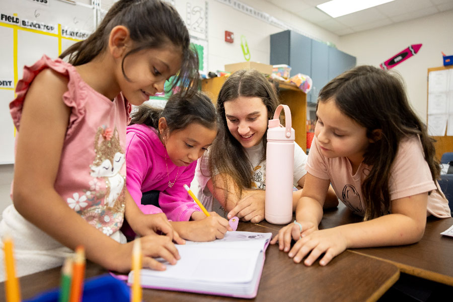 Group of students work together with a teacher at a classroom table, writing and drawing in a notebook