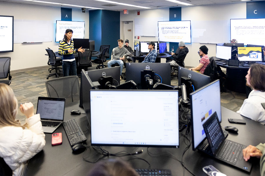 Instructor leads discussion with students in a computer lab classroom with multiple monitors