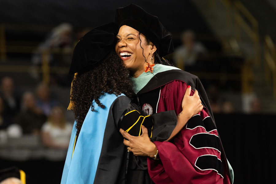 Graduate and faculty member in academic regalia embrace during a graduation ceremony on stage