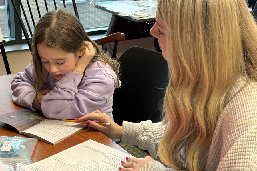 Adult guiding a child reading a book at a table, supporting literacy development in a learning program.