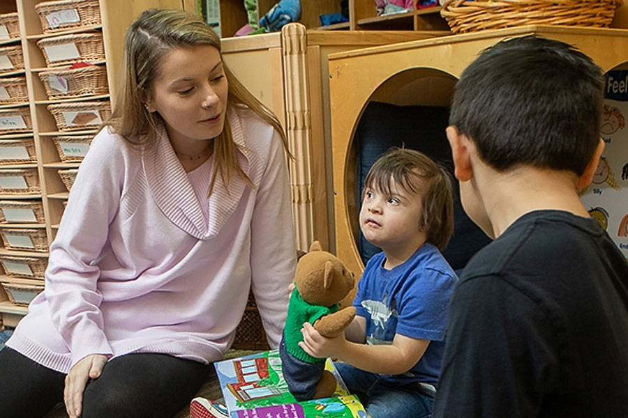 Adult interacting with two children in a classroom, using a plush toy during a learning activity.