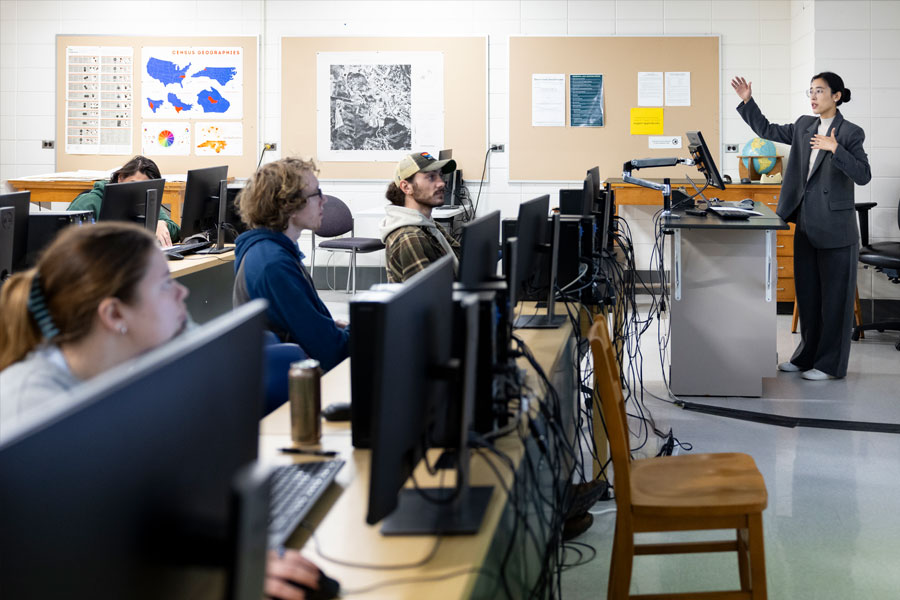 Instructor at front of classroom teaching students working at desktop computers in a lab setting.