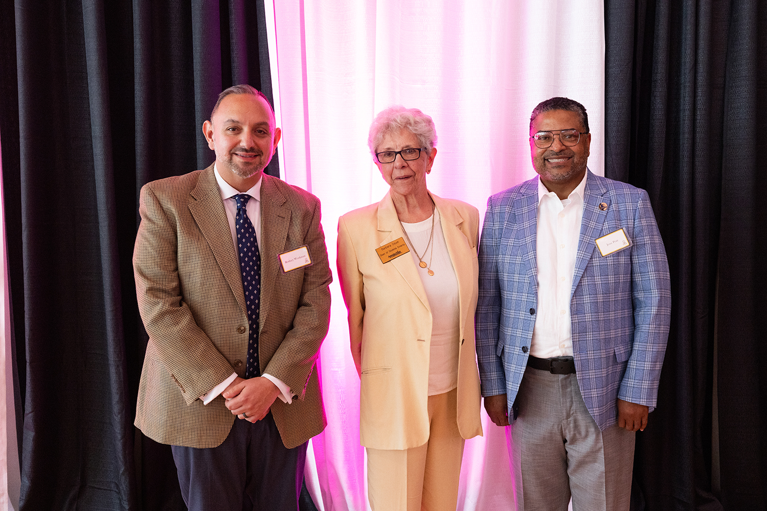 Three individuals in business attire stand posing together in front of white draped curtains with pink lighting at an indoor event.