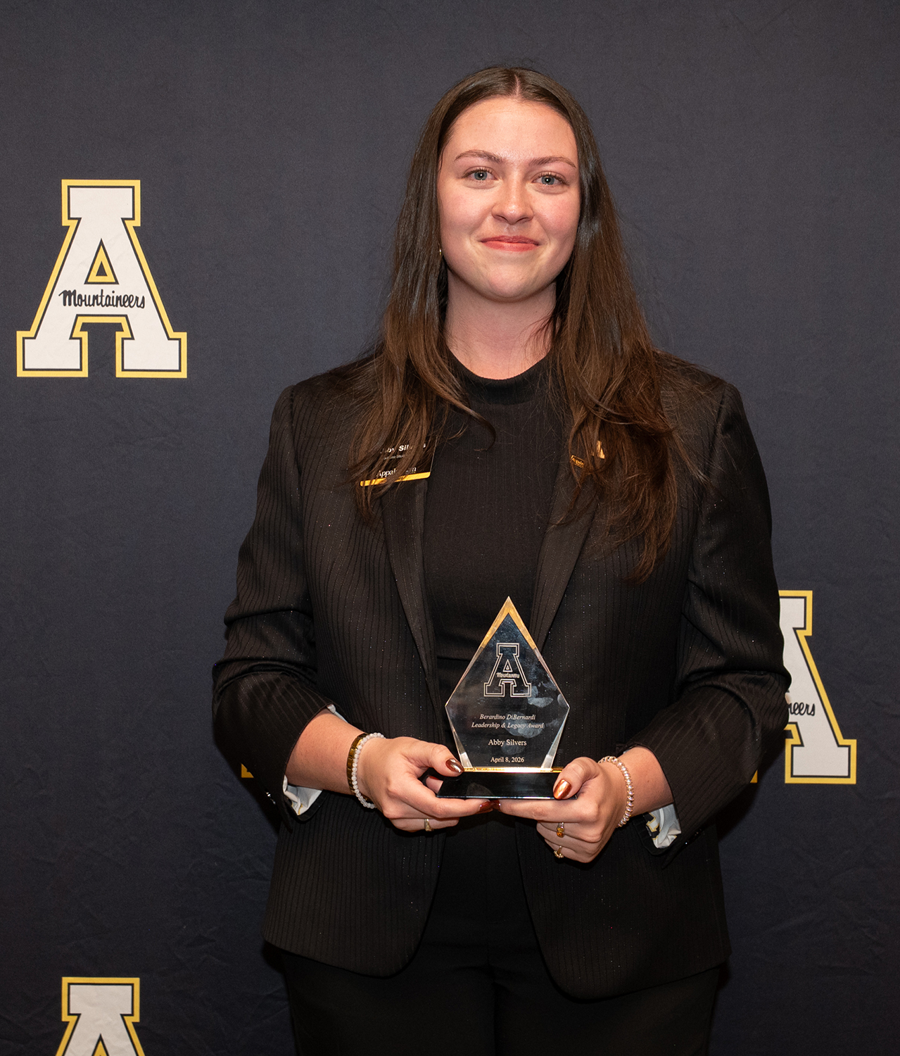 College student in black pant suit holds award.