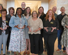 Group of approximately fifteen adults standing indoors in a campus hallway, several holding clear glass awards during a recognition event.