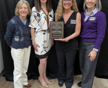 Four people are smiling and standing together indoors in front of a black curtain. Three are upright, and one is crouching in front. The person standing second from the left is holding a plaque. They appear to be dressed in business attire.