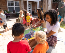 Adult shows an “Animal A-Z” book to two children in an outdoor play area with other kids nearby.