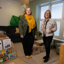 A cozy early‑learning classroom with two adults standing among bookshelves, children’s artwork, a couch, and play materials. The room features natural light from large windows, comfortable seating, and organized activity areas designed for young children.