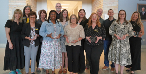 Group of approximately fifteen adults standing indoors in a campus hallway, several holding clear glass awards during a recognition event.