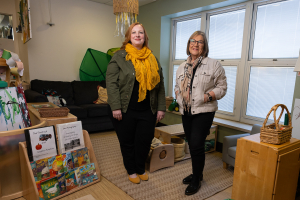 A cozy early‑learning classroom with two adults standing among bookshelves, children’s artwork, a couch, and play materials. The room features natural light from large windows, comfortable seating, and organized activity areas designed for young children.