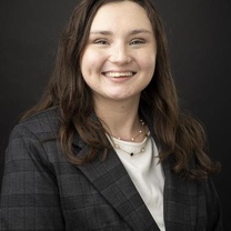 Professional headshot of a person with long dark hair wearing a dark plaid blazer over a light-colored top against a dark background.