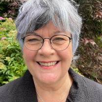 Professional headshot of a person with short gray hair and round glasses, wearing a dark jacket, photographed outdoors with greenery in the background.