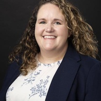 Professional headshot of a person with shoulder-length curly hair wearing a navy blazer over a white floral-patterned blouse against a dark background.