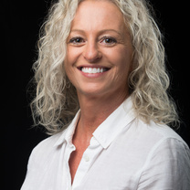 Professional headshot of a person with shoulder-length curly light-colored hair wearing a white button-up shirt against a dark background.