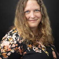 Professional headshot of a person with shoulder-length wavy hair wearing a black floral-patterned top against a dark background.