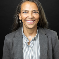 Professional portrait with black background. Person wearing grey blazer and striped shirt and smiling.