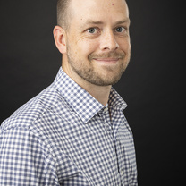 Professional headshot of a person wearing a blue-and-white checkered button-down shirt against a dark background.