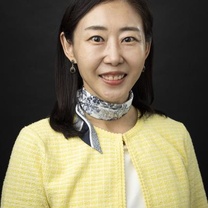 Professional headshot of a person wearing a light yellow jacket and patterned neck scarf against a dark background.
