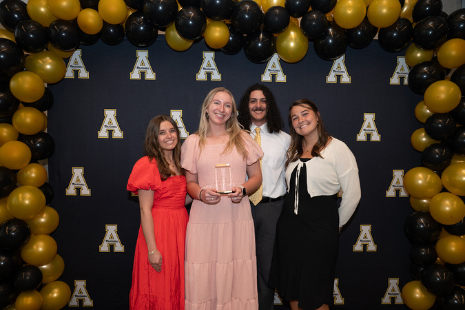 Four people pose with an award beneath black and gold balloons on a backdrop of repeated App State block A logos.