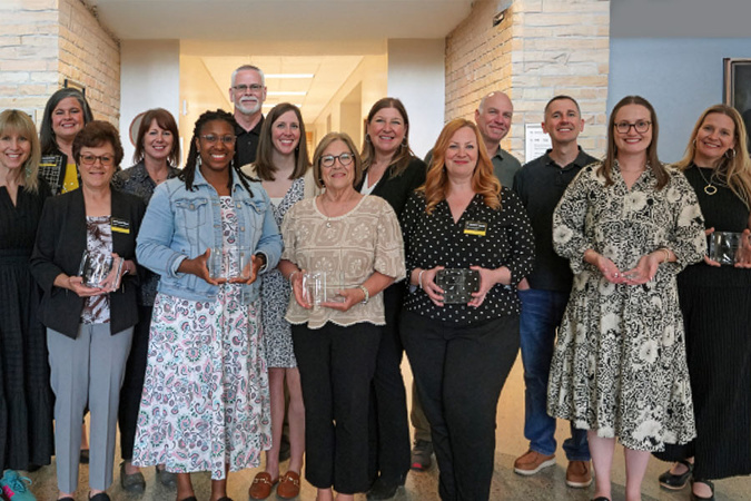 Group of approximately fifteen adults standing indoors in a campus hallway, several holding clear glass awards during a recognition event.