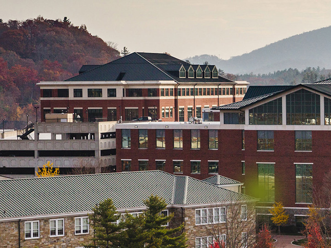 A view of several brick and stone campus buildings with green roofs, set against wooded hills and mountains.