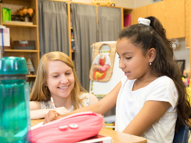 Teacher assisting a student at a classroom desk with a pencil case and school supplies nearby.